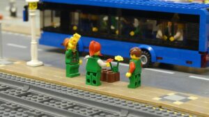 Three Lego people wait on a train platform. A bus full of lego people passes by them.